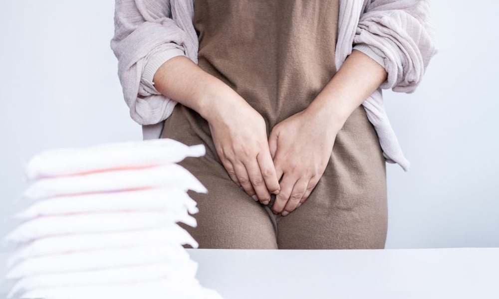 Une femme en gros plan est au centre de l'image. Elle se tient l'entre jambe au niveau du vagin car elle souffre de brûlures vulvaires à cause de la ménopause. Devant elle se trouve ses serviettes hygiéniques.