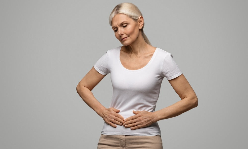 Une femme ménopausée est au centre de l'image. Elle se tient le ventre avec ses deux mains car elle a une douleur en bas du ventre à cause de la ménopause. Elle fait une grimace et a le regard vide. Le fond est gris.