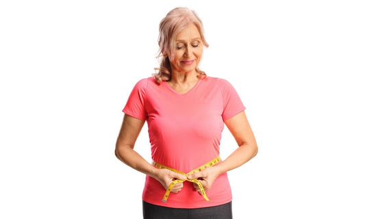 Une femme qui a la ménopause et qui subit une perte de poids est au centre de l'image. Elle sourit car elle se sent mieux. Elle mesure son tour de ventre avec un mètre jaune. Son T-shirt est rose et le fond blanc.