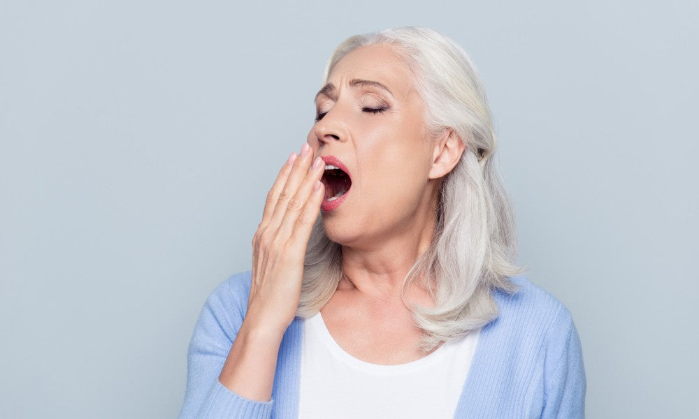 Une femme d'âge moyen aux cheveux blancs est au centre de l'image. Elle baye et tente de mettre sa main droite devant sa bouche. Elle subit une fatigue extrême à cause de la ménopause. Son gilet est bleu, et le fond d'un bleu un peu plus clair.