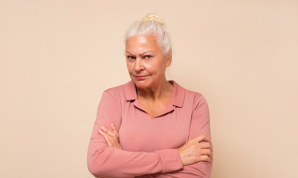 Une femme âgée de 50 ans qui souffre de la ménopause et de troubles de l'humeur est au centre de l'image. Elle croise le bras car elle énervée et ne trouve pas de traitement pour se calmer.
