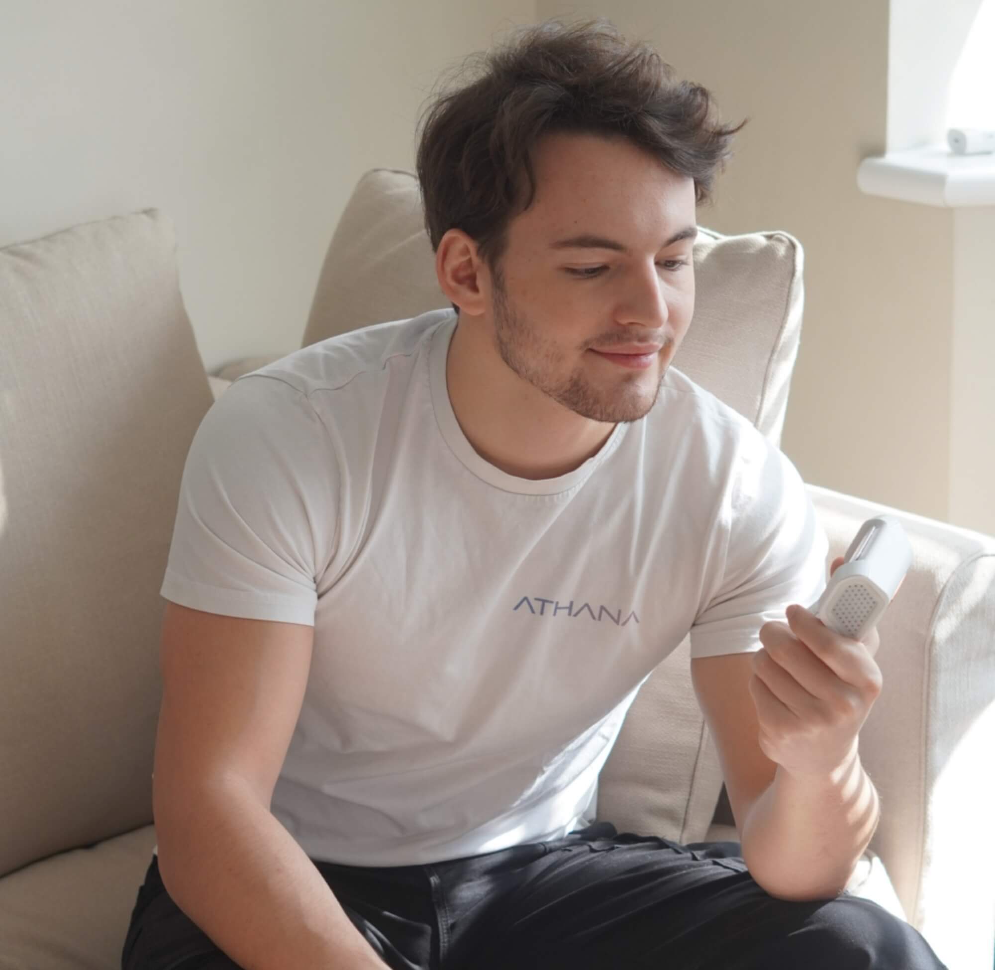 Un homme assis sur un canapé, portant un t-shirt blanc floqué "ATHANA", tient dans sa main le dispositif Héra et le regarde avec satisfaction. L’image illustre l’implication personnelle du fondateur dans le développement du produit.