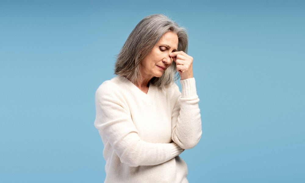 Une femme mûre d'à peu près 50 ans est au centre de l'image en gros plan. Elle se tient la tête avec la main gauche, car elle est en dépression à cause de la ménopause. Elle porte un pull blanc et le fond est bleu.
